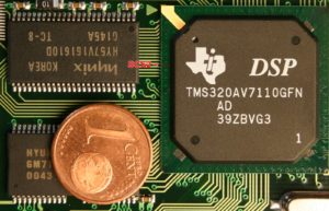 Abbildung 5: Nur unter der Lupe lassen sich die feinen Leiterbahnen am Rand des MPEG-Decoders erahnen, der Pfeil zeigt auf das ebenfalls rot markierte SDRAM Chip Select 2. Zum Größenvergleich liegt links unten ein 1-Cent-Stück.
